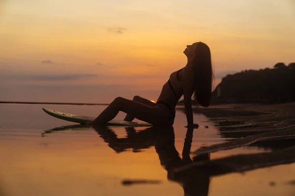 Silhouette sexy girl in swimsuit lying and posing on surf board at beach during sunset — Stock Photo, Image