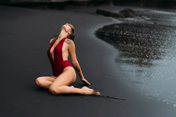 Sexy Model mit großen Brüsten im roten Badeanzug posiert am schwarzen Sandstrand. — Stockfoto