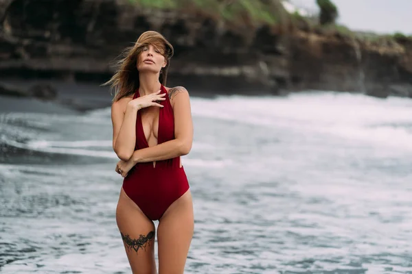 Сексуальна модель з великими грудьми в червоному купальнику позує на чорному піщаному пляжі . — стокове фото