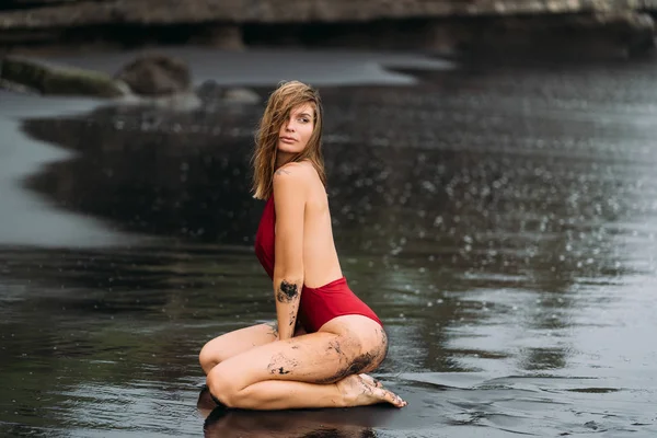 Sexy model with big breasts in a red swimsuit poses on black sand beach. — Stock Photo, Image