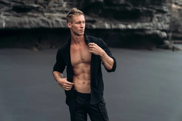 Portrait of handsome muscular man in black shirt posing on camera at black sandy beach
