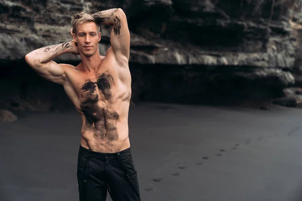 Sexy fitness male model in black pants and shirtless posing on black sandy beach