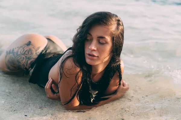 Portrait of sexy brunette girl in bikini and knitted sweater lies on the sandy beach, tattoo on her buttocks. — Stock Photo, Image