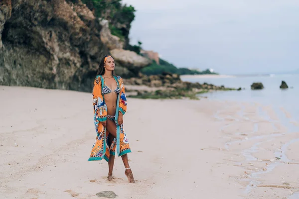 Hermosa chica sexy en traje de baño caminando en la playa de arena con roca en el fondo — Foto de Stock