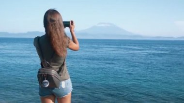Sırt çantası ile kız gezgin arka yan görünümü telefonda okyanus ve volkan fotoğraf çeker