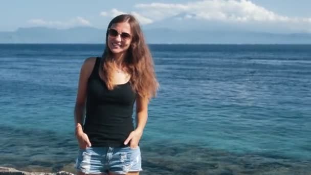 Beautiful girl traveler in sunglasses stands near ocean and poses on camera — Stock Video