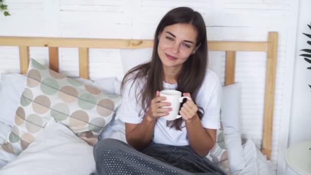 Hermosa chica se sienta en la cama, sostiene la taza, mira a la cámara y sonríe, cámara lenta — Vídeos de Stock