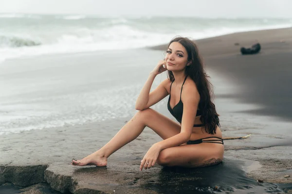 Beau modèle brune en maillot de bain assis sur une plage de sable noir . — Photo