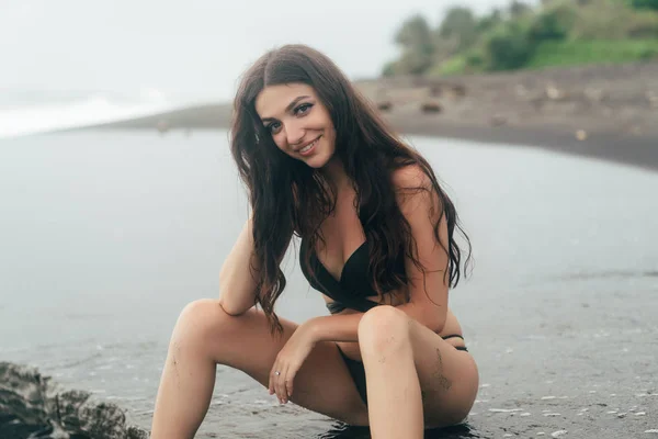 Mooie brunette model in zwembroek zittend op zwart zandstrand. — Stockfoto