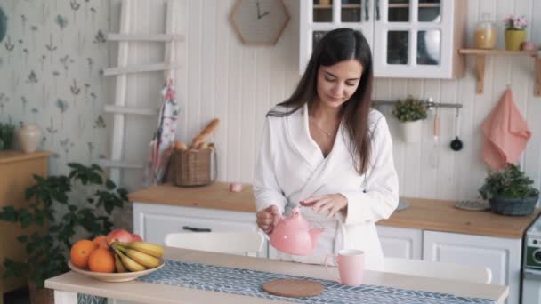 Girl in white bathrobe at home kitchen pours tea in cup in morning, slow motion — Stock Video