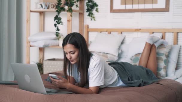 Linda chica freelancer se encuentra en la cama y utiliza el teléfono y el ordenador portátil para el trabajo, cámara lenta — Vídeos de Stock