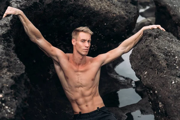 Portrait de jeune homme avec torse nu debout sur la plage de roche avec du sable noir — Photo