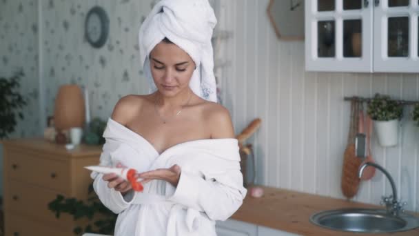 Красивая девушка в халате, с полотенцем на голове наносит крем на тело, замедленное движение — стоковое видео