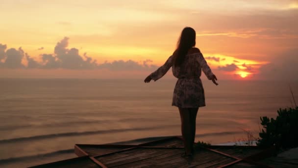 Menina estende os braços bem abertos na praia olha para o pôr-do-sol e oceano, câmera lenta — Vídeo de Stock
