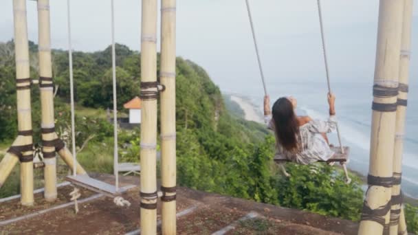 Vista trasera de la joven mujer columpios en columpio con hermosa vista sobre el océano, cámara lenta — Vídeo de stock