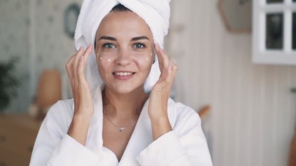 Närbild, flicka i badrock, med handduk på huvudet applicerar grädde på hennes ansikte, slow motion — Stockvideo