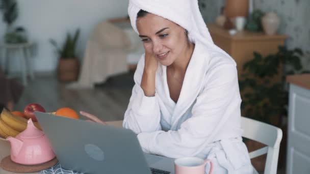 Vrouw in witte badjas, met handdoek op hoofd kijkt in laptop, glimlacht, slow motion — Stockvideo