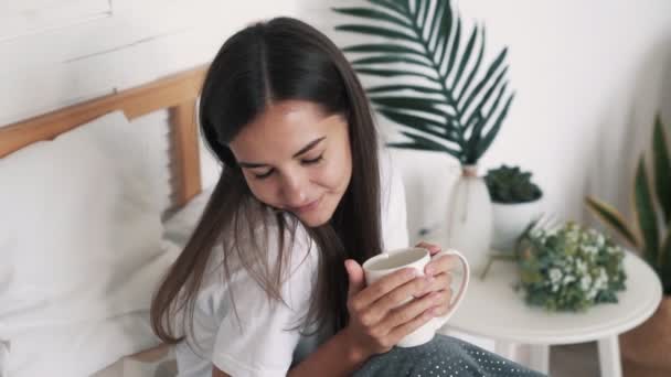 Hermosa chica se sienta en la cama, sostiene la taza, disfruta de la mañana y sonríe, cámara lenta — Vídeos de Stock