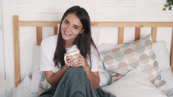 Chica envuelta en manta se sienta en la cama sostiene taza, mira a la cámara, sonríe, cámara lenta — Vídeos de Stock