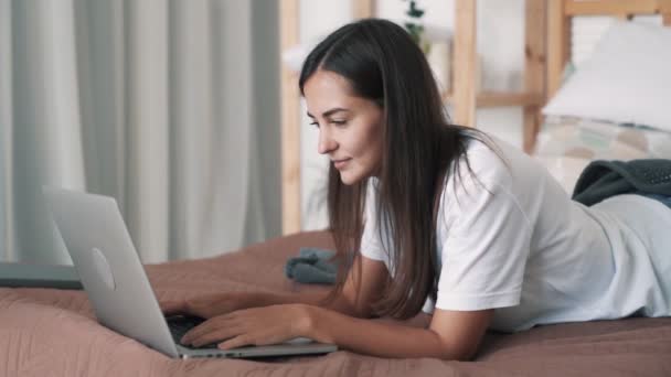 Portré lány fekszik az ágyban a gyomrán, típusok laptop billentyűzeten, lassított felvétel — Stock videók