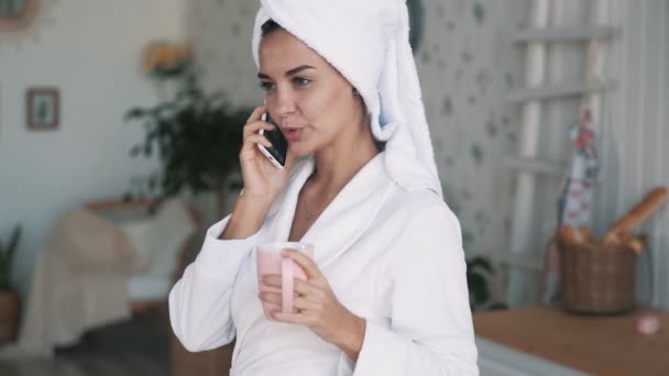Девушка с полотенцем на голове, разговаривает по телефону, держит чашку в руках, замедленная съемка — стоковое видео
