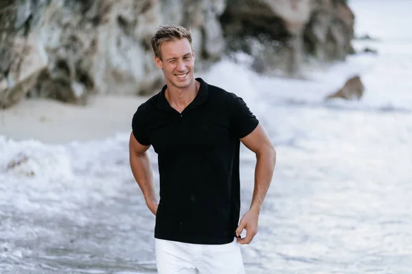 해변에서 휴식 검은 티셔츠에 섹시한 웃는 남자, 배경에 바다 파도. — 스톡 사진