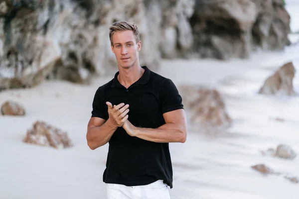 Sexy muscular man in black t-shirt resting on beach, ocean waves at background.