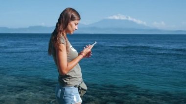Kız gezgin telefon kullanır, mesaj türleri. Okyanus, arka planda dağlar, yavaş çekim