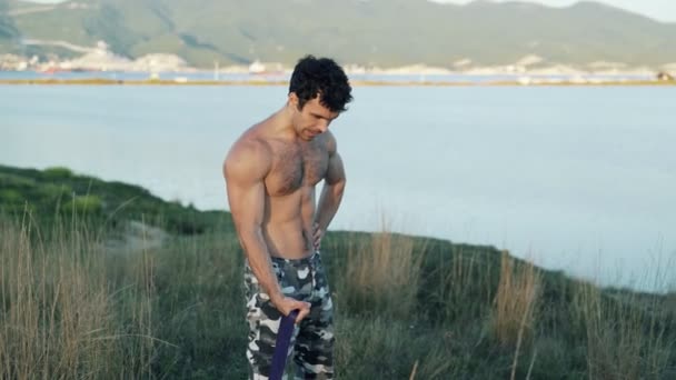 Atleta haciendo ejercicios con una banda elástica de fitness sobre un fondo de montañas y el mar . — Vídeo de stock