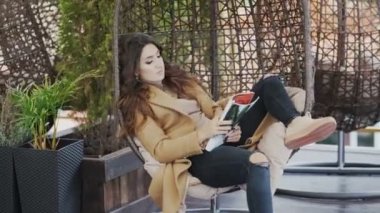 Kazak ve paltolu güzel genç kadın kafede sandalyede oturur ve dergi okur