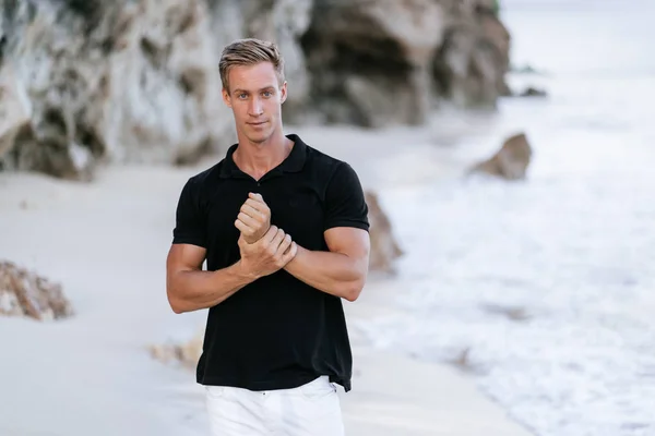 Sexy muscular man in black t-shirt resting on beach, ocean waves at background.