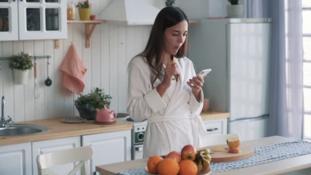 Mujer joven en albornoz en la cocina come pedazo de manzana y utiliza el teléfono, cámara lenta — Vídeo de stock