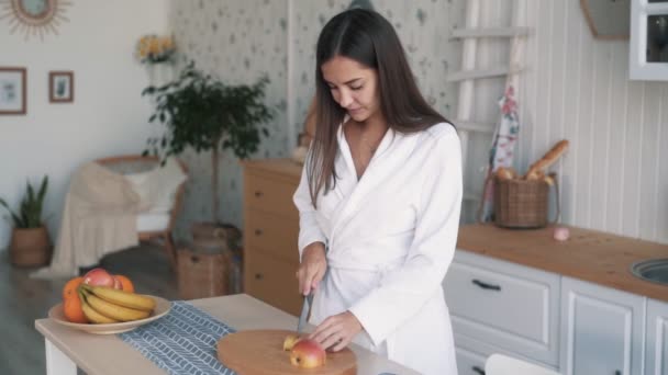 Cute Girl w białym szlafroku tnie jabłko na pokładzie w kuchni, w zwolnionym tempie — Wideo stockowe