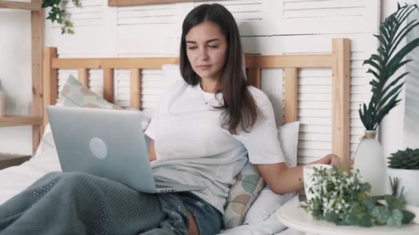 Young woman sits on bed, drinks coffee and works on laptop at bedroom, slow motion — Stock Video