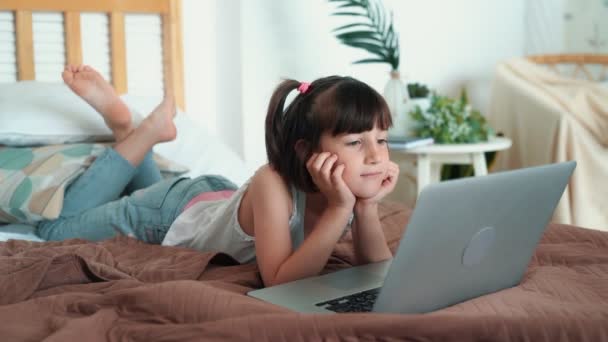 Маленькая девочка лежит на кровати и смотрит мультик на ноутбуке в своей комнате, замедленная съемка — стоковое видео