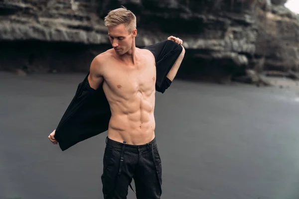 Sexy fitness male model in black pants and shirtless posing on black sandy beach.