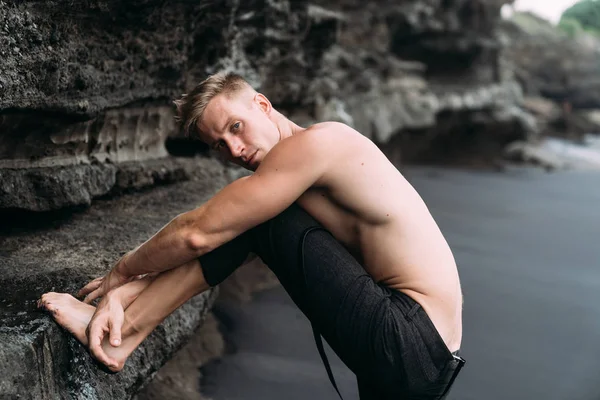 Modèle masculin de fitness sexy en pantalon noir et torse nu posant sur une plage de sable noir . — Photo