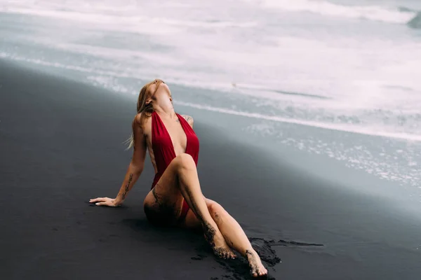 Modelo sexy con grandes pechos en un traje de baño rojo posa en la playa de arena negra . —  Fotos de Stock