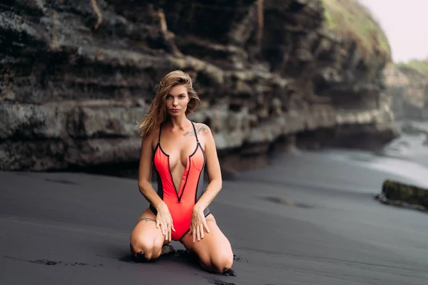 Beautiful sexy girl with big breasts in a red swimsuit sunbathing on black sand beach. — Stock Photo, Image
