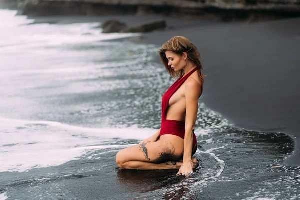 Modelo sexy com seios grandes em um maiô vermelho posa na praia de areia preta . — Fotografia de Stock