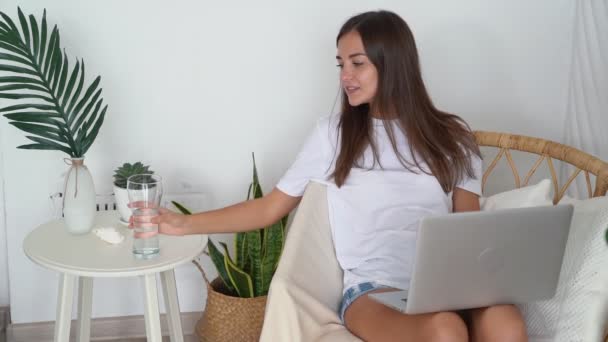 Chica se sienta en la silla con el ordenador portátil en casa, bebe agua clara de vidrio, cámara lenta — Vídeos de Stock