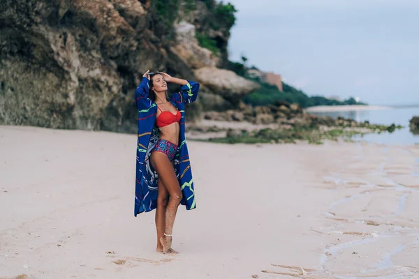 Hermosa chica sexy en traje de baño caminando en la playa de arena con roca en el fondo —  Fotos de Stock