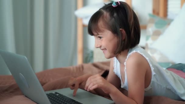 Маленькая девочка лежит на кровати, использует ноутбук, играет в компьютерные игры, замедленная съемка — стоковое видео