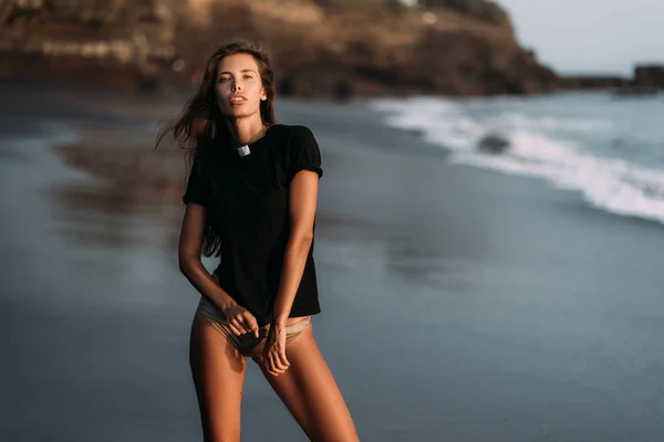 Sexy ragazza bruna in t-shirt e costume da bagno in posa sulla spiaggia con sabbia nera — Foto Stock