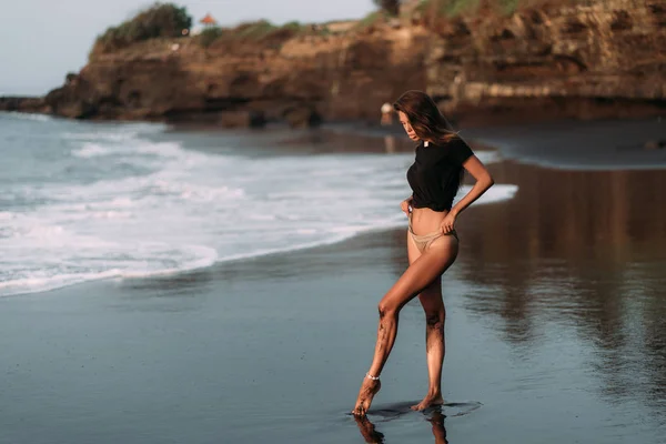 Dívka v černém tričku a plavky na černé písečné pláži v paprscích západu slunce — Stock fotografie