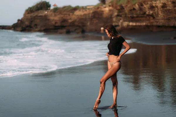 Girl in black T-shirt and swimsuit on black sandy beach in rays of sunset Stock Photo