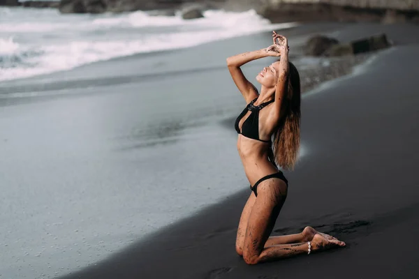 Siyah mayo giyen genç seksi kız siyah volkanik kumsalda dinleniyor. — Stok fotoğraf