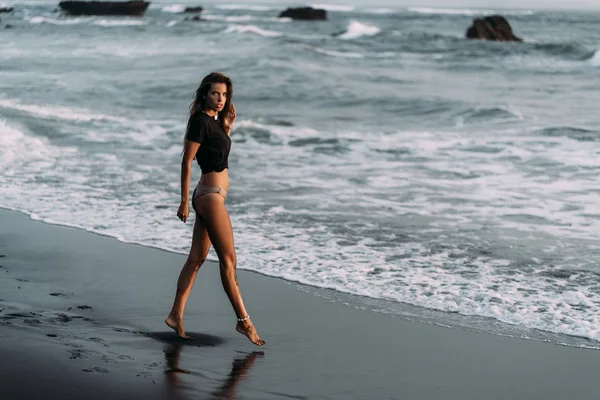 Chica con cuerpo sexy en camiseta negra y trajes de baño camina en la playa de arena negra —  Fotos de Stock