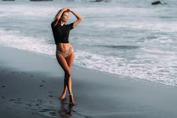 Chica en camiseta negra y traje de baño en la playa de arena negra en los rayos de la puesta del sol — Foto de Stock