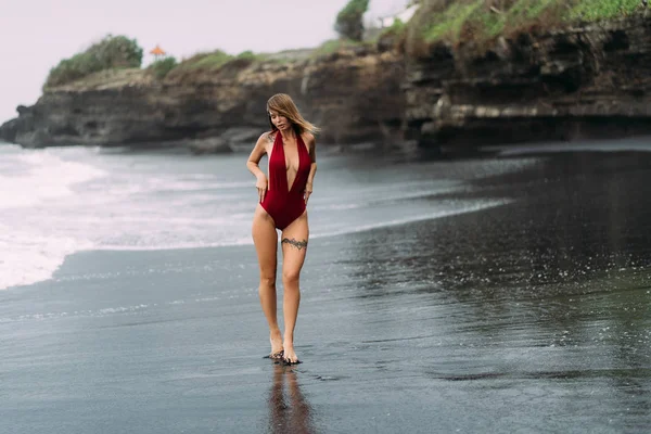 Hermosa chica sexy con grandes pechos en un traje de baño rojo tomando el sol en la playa de arena negra . — Foto de Stock